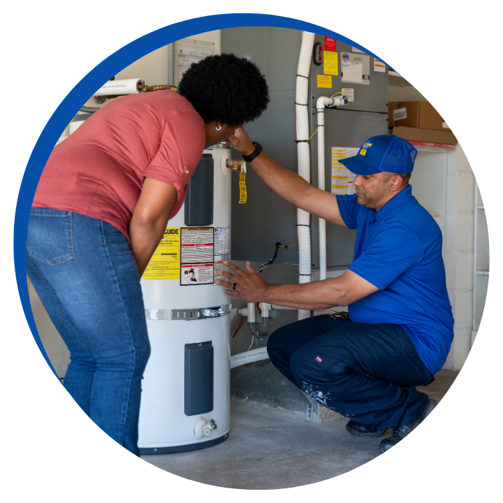 Technician in a blue uniform repairing a water heater and explaining to a customer in a utility room.