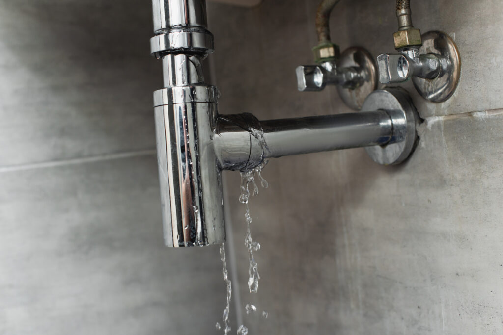 Water leaking from chrome sink pipes on gray tile wall.